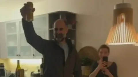 Emil Flatz podiže čašu dok ga Daria Lorenci Flatz snima mobitelom u kuhinji.