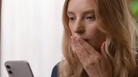 Mlada žena drži ruku preko usta dok iznenađeno gleda u zaslon mobitela.