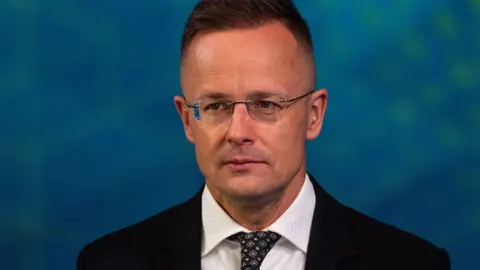 Portret muškarca u tamnom odijelu s naočalama ispred plave pozadine.