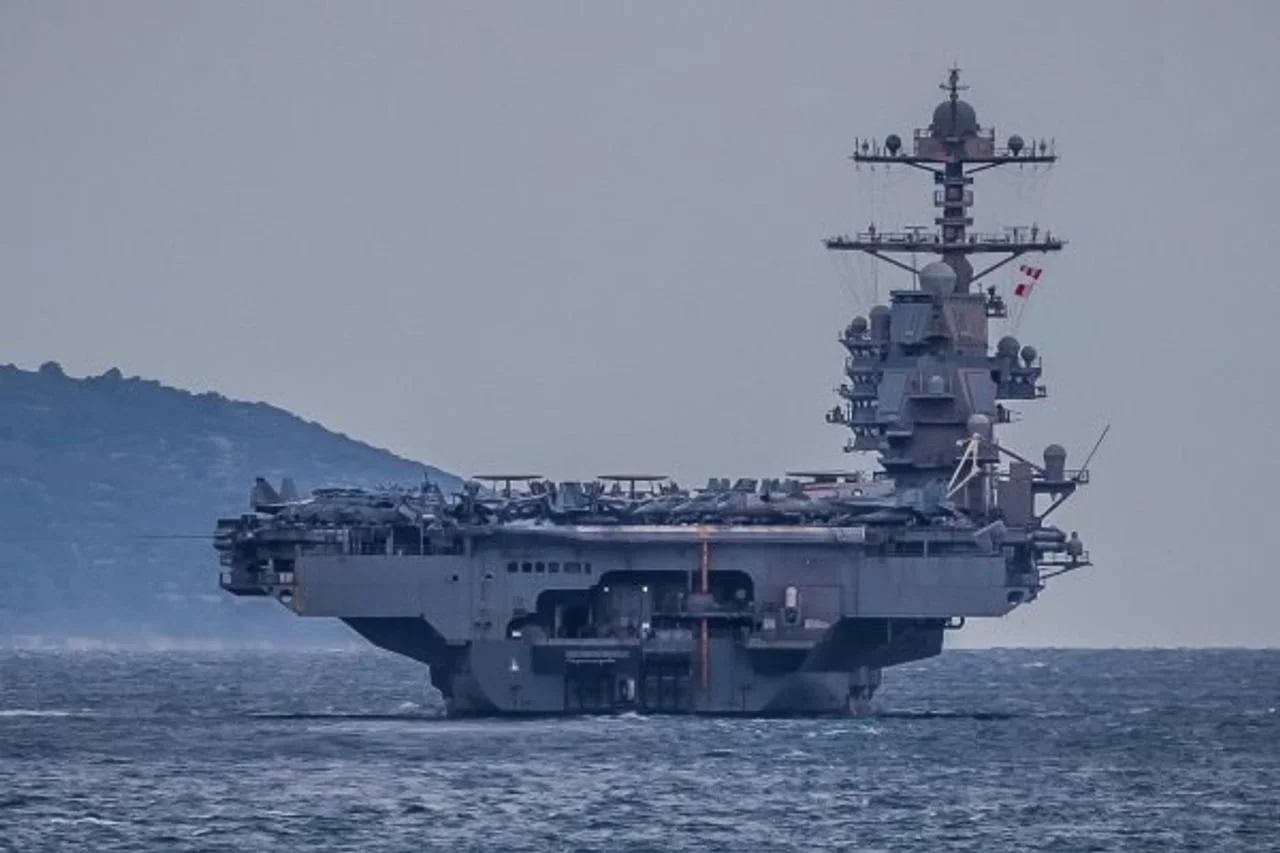 Mornar u crvenoj uniformi i zaštitnom kacigom radi s vojnom opremom na palubi.