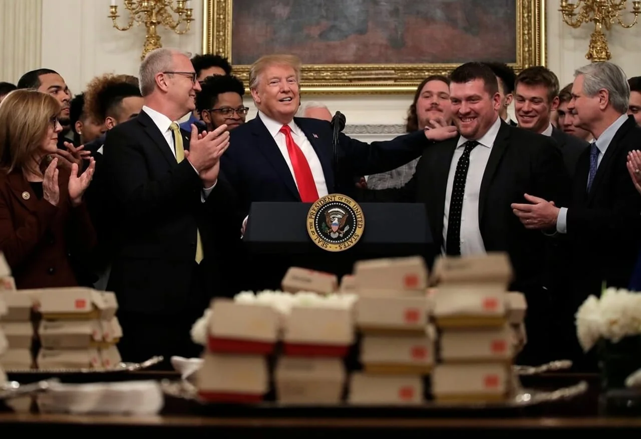 Predsjednik Trump stoji za govornicom okružen ljudima i brzim obrocima