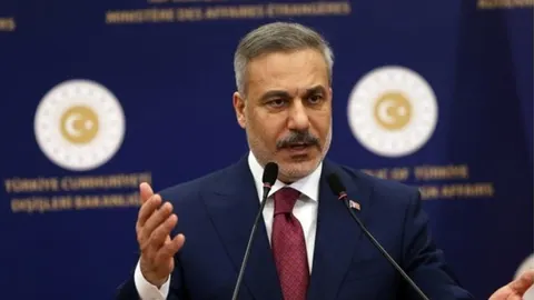 Hakan Fidan govori za govornicom s mikrofonima ispred turskih simbola.