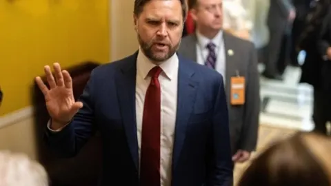 J.D. Vance u tamnom odijelu s crvenom kravatom podiže ruku tijekom govora.