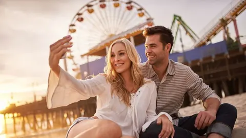 Nasmijani par snima selfie na plaži ispred zabavnog parka tijekom zalaska sunca.