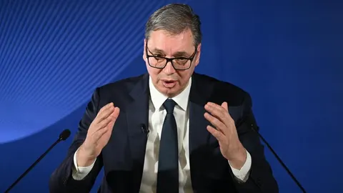 Aleksandar Vučić govori za govornicom s podignutim rukama na plavoj pozadini.