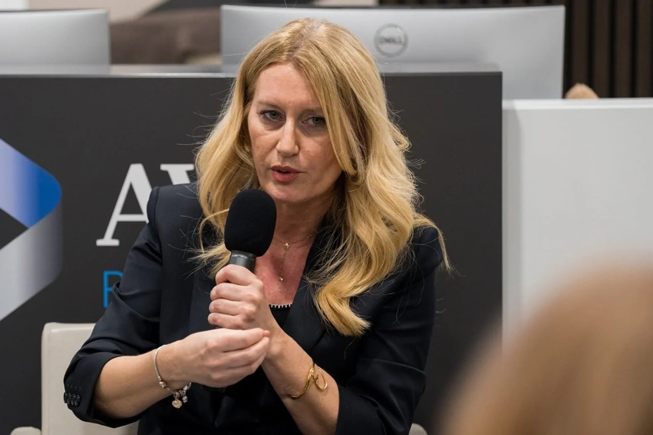 Plavokosa žena u tamnom sakou objašnjava temu držeći mikrofon objema rukama.