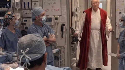 Donald Trump u bijeloj halji i crvenom plaštu ulazi u bolničku operacijsku salu.
