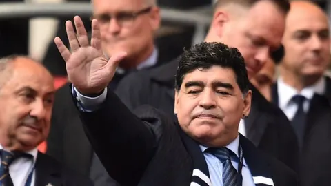 Diego Maradona u odijelu maše rukom publici na stadionu.