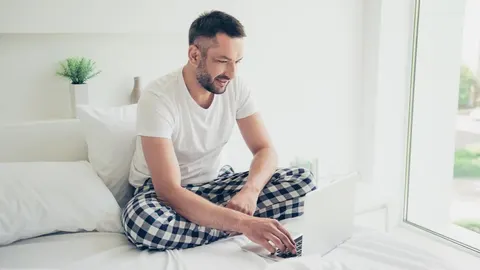 Muškarac u pidžami sjedi na krevetu i koristi prijenosno računalo