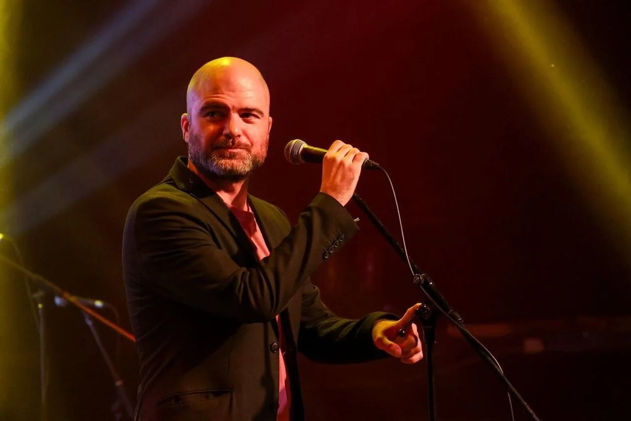 Filip Riđički s obrijanom glavom nastupa u crnom sakou pod reflektorima.