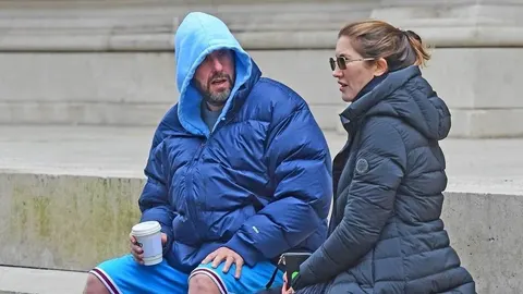 Adam Sandler u plavoj zimskoj jakni i kratkim hlačama sjedi vani