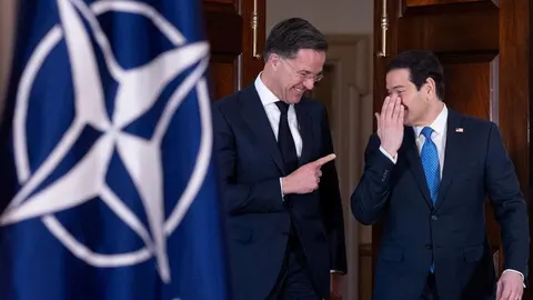 Dva muškarca u odijelima razgovaraju ispred NATO zastave.