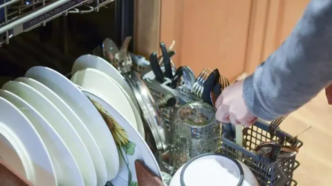 Ruka stavlja prljavi pribor za jelo u košaru perilice posuđa uz tanjure.