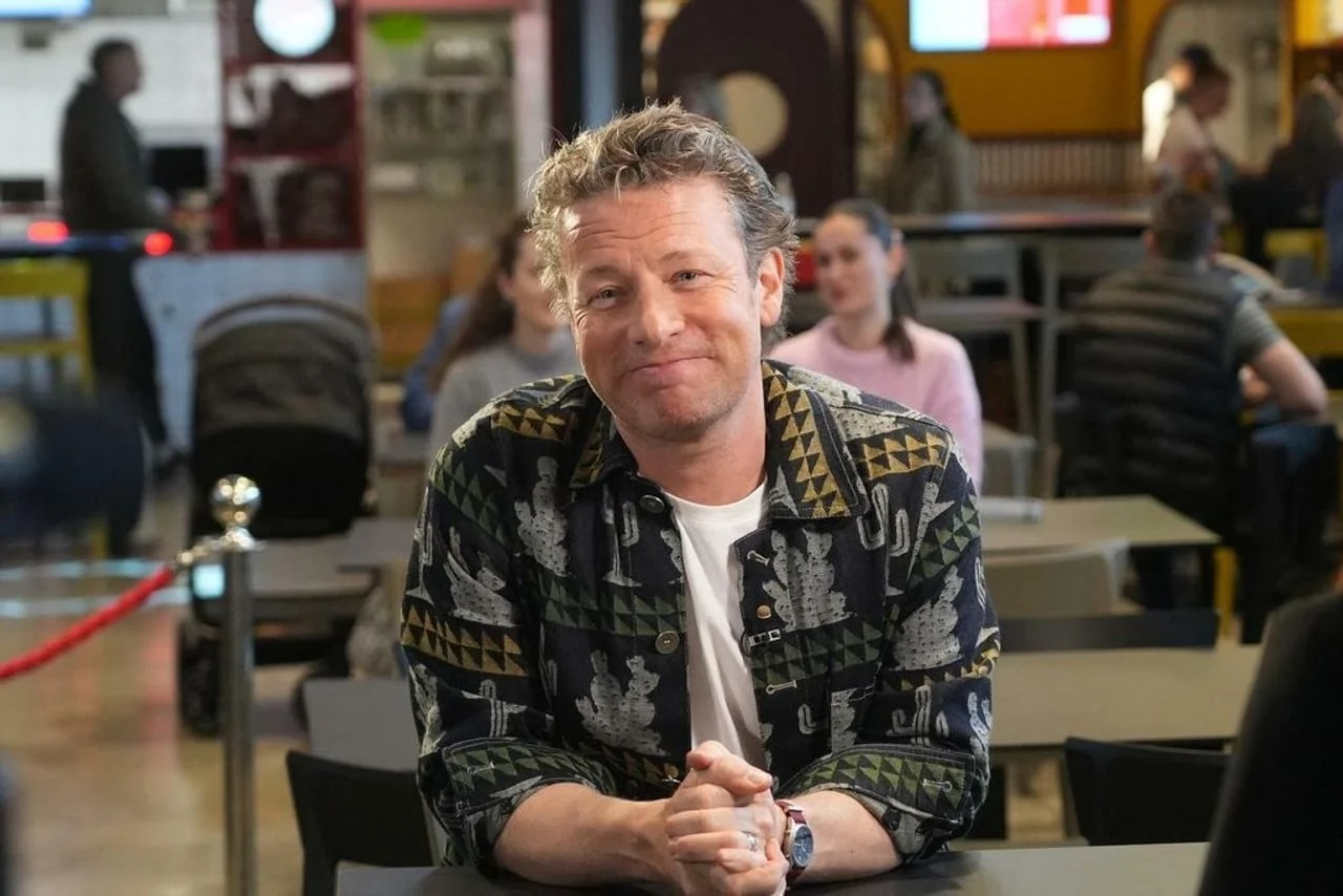 Nasmiješeni Jamie Oliver sjedi za stolom s prekriženim rukama u opuštenom izdanju.