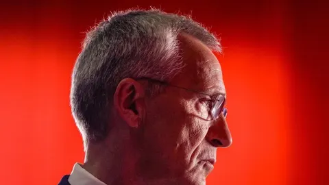 Profil Jensa Stoltenberga s naočalama ispred jarko crvene pozadine.