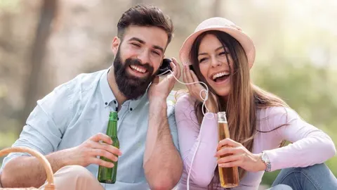 Nasmijani par na pikniku pije piće i sluša glazbu na slušalicama.