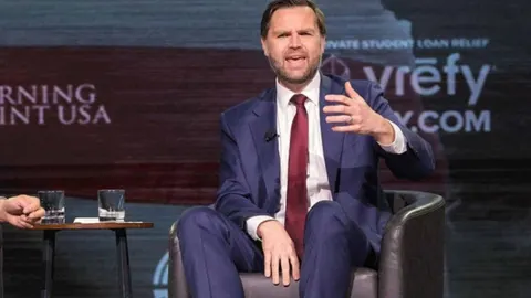 JD Vance u odijelu s crvenom kravatom govori na pozornici dok gestikulira rukom.