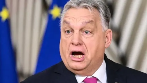 Viktor Orbán u krupnom planu dok govori s otvorenim ustima ispred zastava EU.