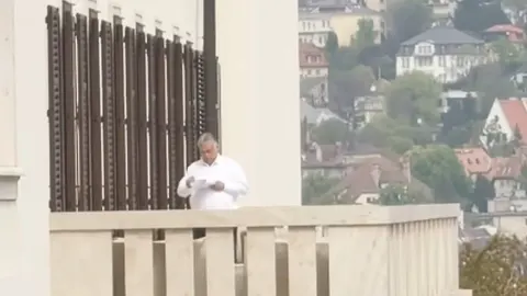 Viktor Orbán stoji na balkonu u bijeloj košulji i čita dokumente.