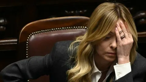 Krupni kadar Giorgie Meloni koja rukom prekriva lice dok sjedi u kožnom naslonjaču.