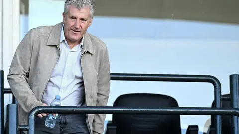 Davor Šuker u svijetloj jakni i košulji drži bocu vode na tribinama.