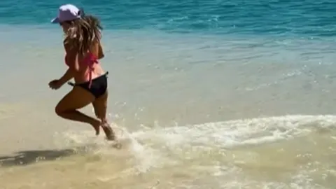 Žena u kupaćem kostimu trči kroz plićak pješčane plaže prema moru.