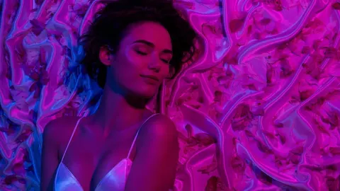 Žena zatvorenih očiju leži na ružičastoj svili okružena laticama cvijeća.