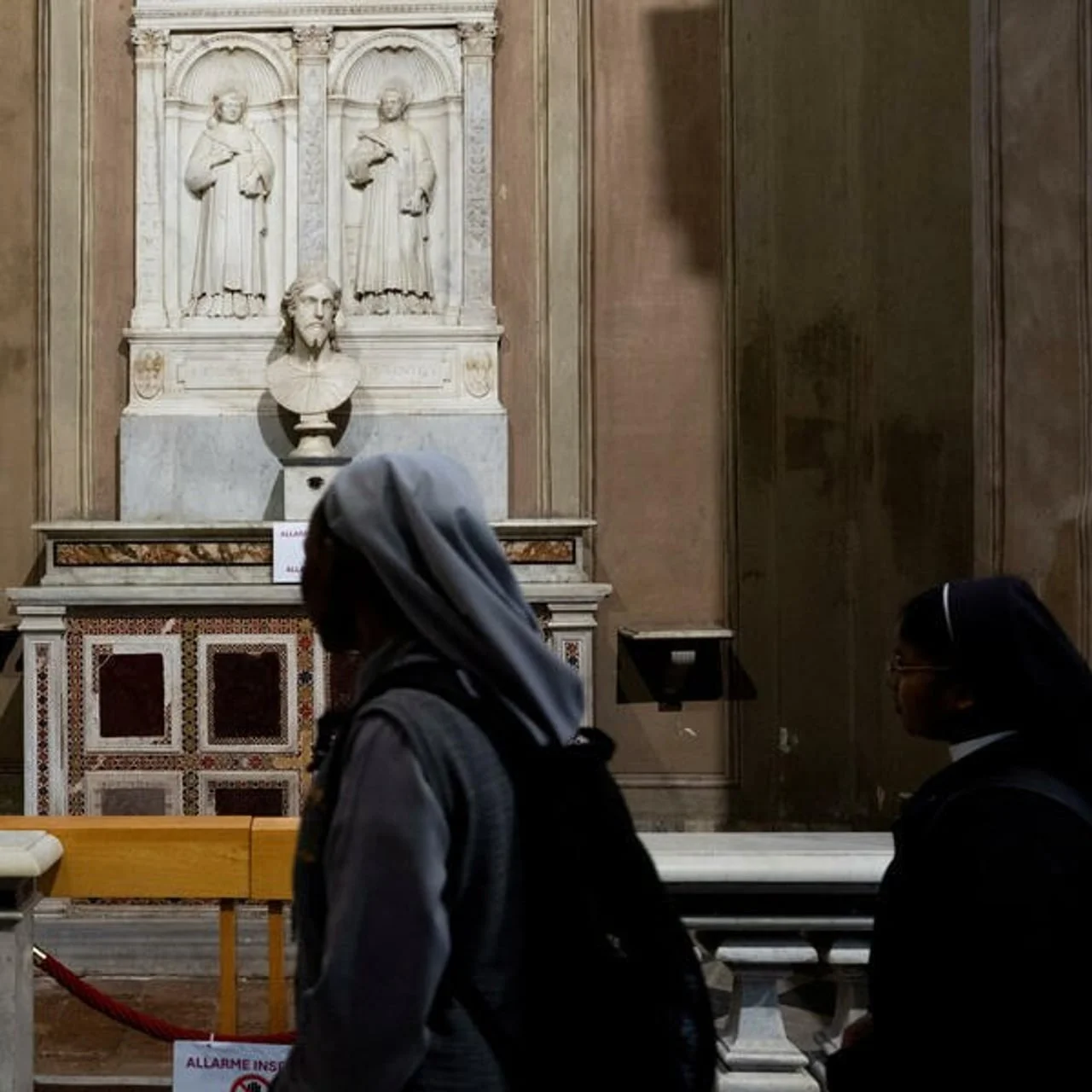 Dvije redovnice u prolazu ispred osvijetljenog oltara s bistom.