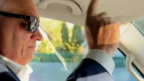 Viktor Orban sa sunčanim naočalama vozi automobil i gestikulira rukom.