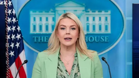 Glasnogovornica Bijele kuće govori za govornicom ispred plave pozadine s logotipom.