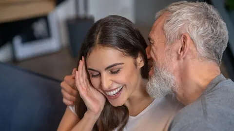 Stariji muškarac sijede brade šapće mladoj nasmijanoj ženi u interijeru.