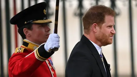Princ Harry u tamnom odijelu prolazi pored kraljevske straže u crvenoj odori