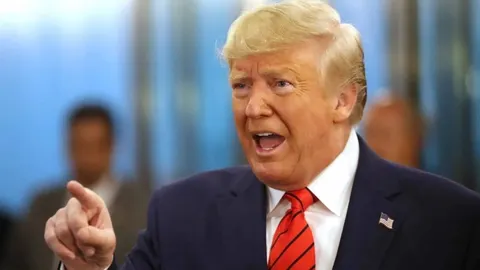 Donald Trump u tamnom odijelu s crvenom kravatom govori i pokazuje prstom