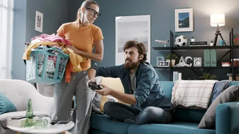 Žena drži košaru s rubljem dok muškarac sjedi na kauču i igra videoigre.