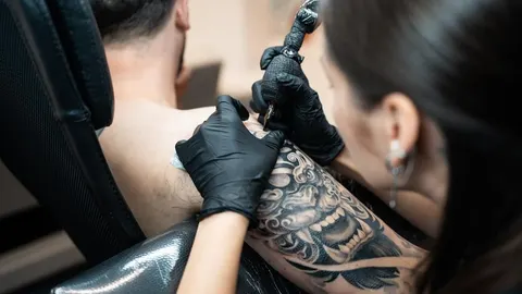 Tattoo majstor s crnim rukavicama nanosi tintu na ruku klijenta pomoću igle.