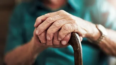Krupni plan naboranih ruku starije osobe koje se oslanjaju na drveni štap za hodanje.