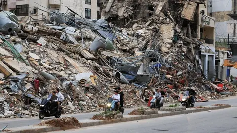 Srušena zgrada i hrpa betonskih ruševina uz cestu kojom prolaze motociklisti.