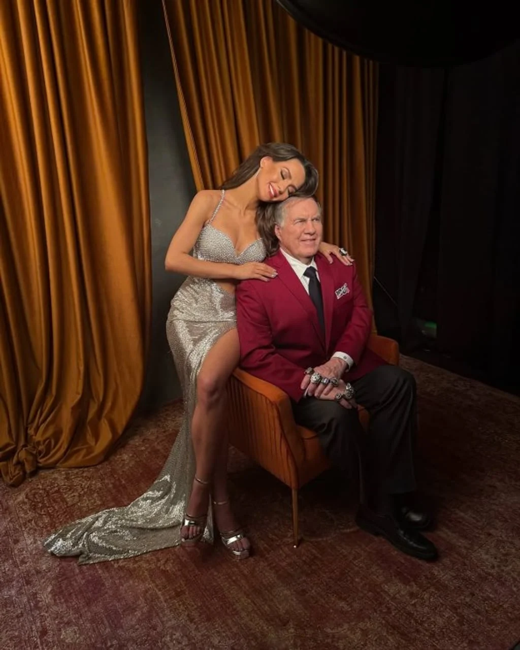 Bill Belichick u crvenom sakou sjedi dok Jordon Hudson u elegantnoj haljini stoji uz njega.