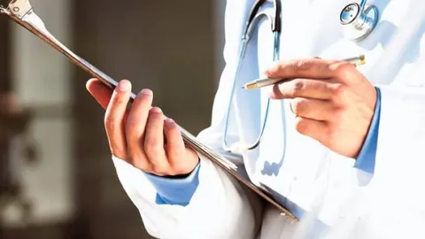 Liječnik u bijeloj kuti drži medicinsku dokumentaciju i olovku.