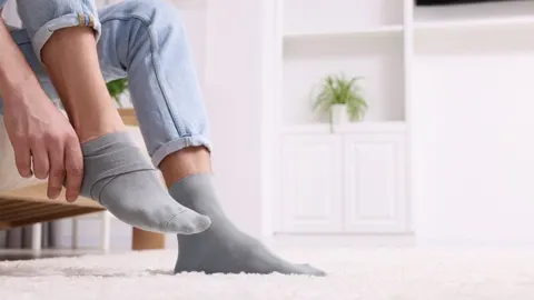 Osoba sjedi i navlači sivu čarapu na stopalo dok nosi traperice.