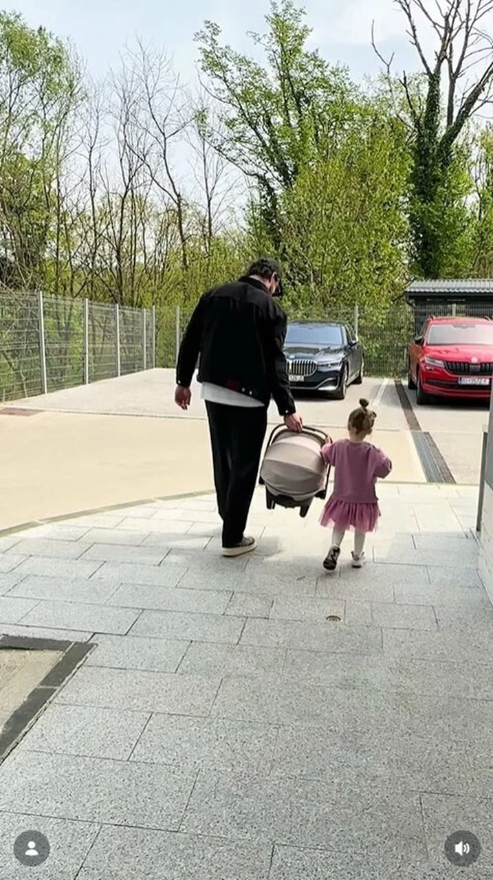 Muškarac nosi autosjedalicu s bebom dok malena djevojčica hoda pored njega na parkingu.