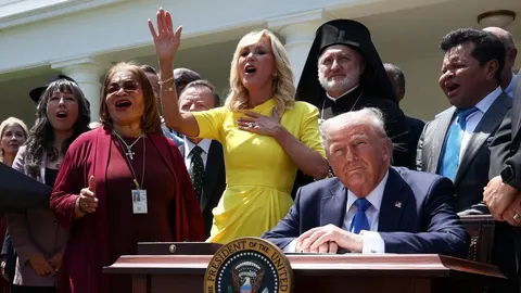 Donald Trump sjedi za govornicom dok skupina ljudi pjeva iza njega na otvorenom.