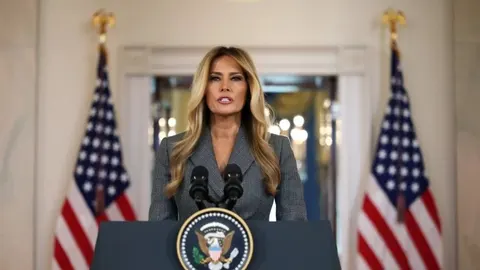 Melania Trump stoji za govornicom s američkim zastavama u pozadini.