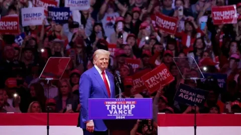 Donald Trump stoji na pozornici ispred mnoštva pristaša s transparentima.