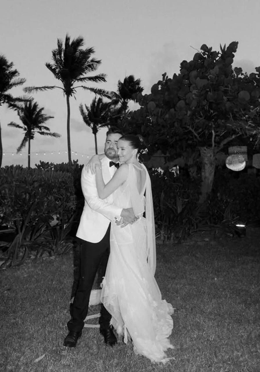 Mladenci u zagrljaju na otvorenom s palmama u pozadini, crno-bijeli prikaz.