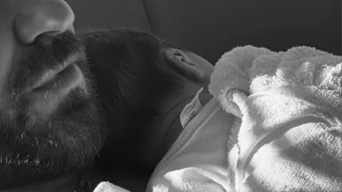 Crno-bijela fotografija bradatog muškarca koji drži novorođenče na prsima.