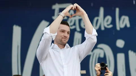 Jordan Bardella u bijeloj košulji rukama oblikuje srce ispred plave pozadine.