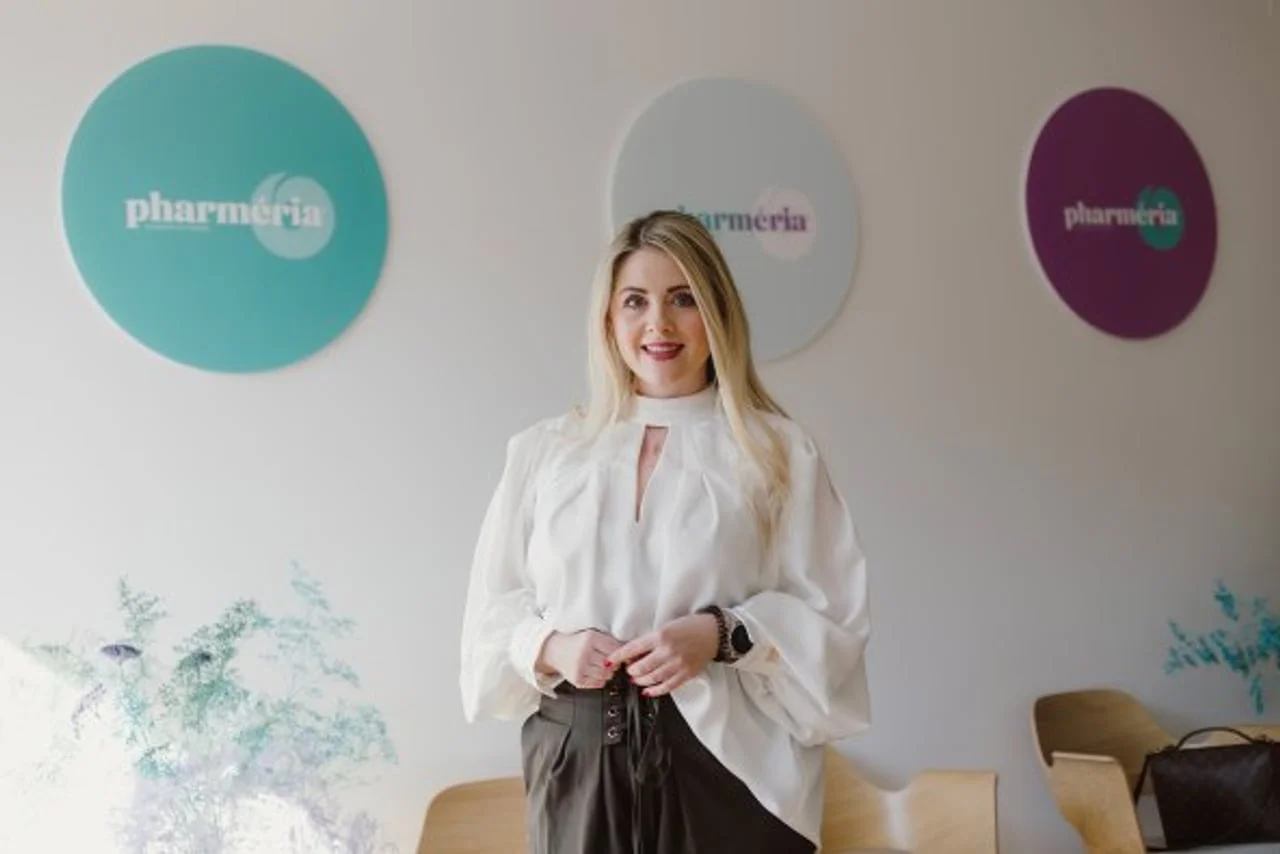 Plavokosa žena u bijeloj bluzi pozira ispred kružnih dekoracija na zidu.