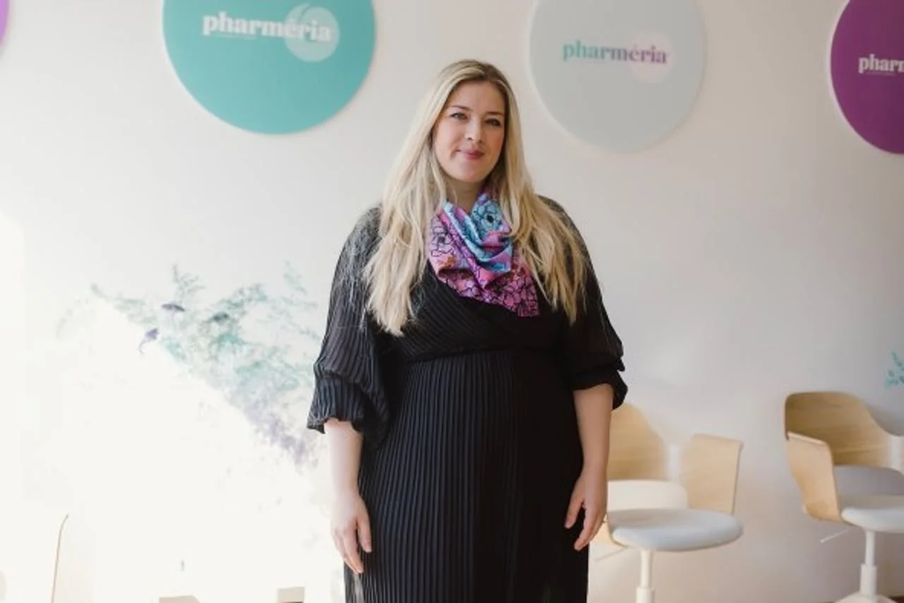 Žena u crnoj odjeći s plavo-ružičastom maramom stoji u svijetloj prostoriji.