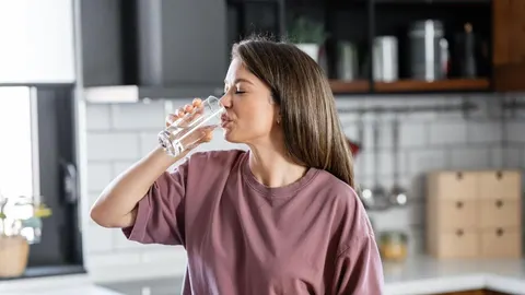 Mlada žena pije svježu gaziranu vodu iz prozirne čaše u kuhinji.
